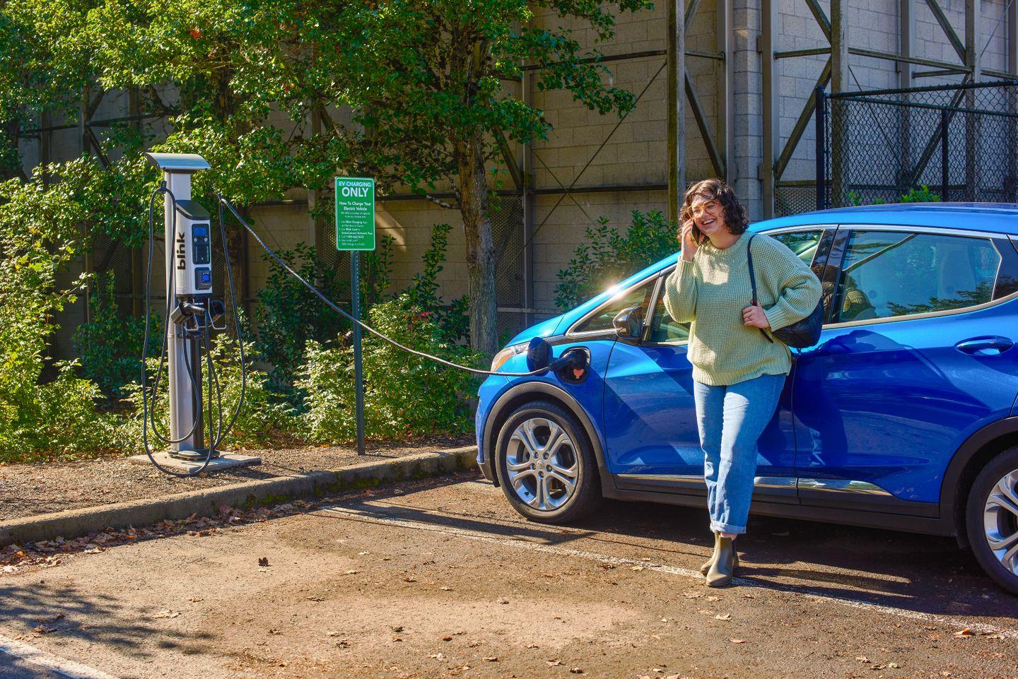 Woman in a green sweater laughs on the phone as her blue electric car charges at an outdoor Blink Series 8 EV charging station.